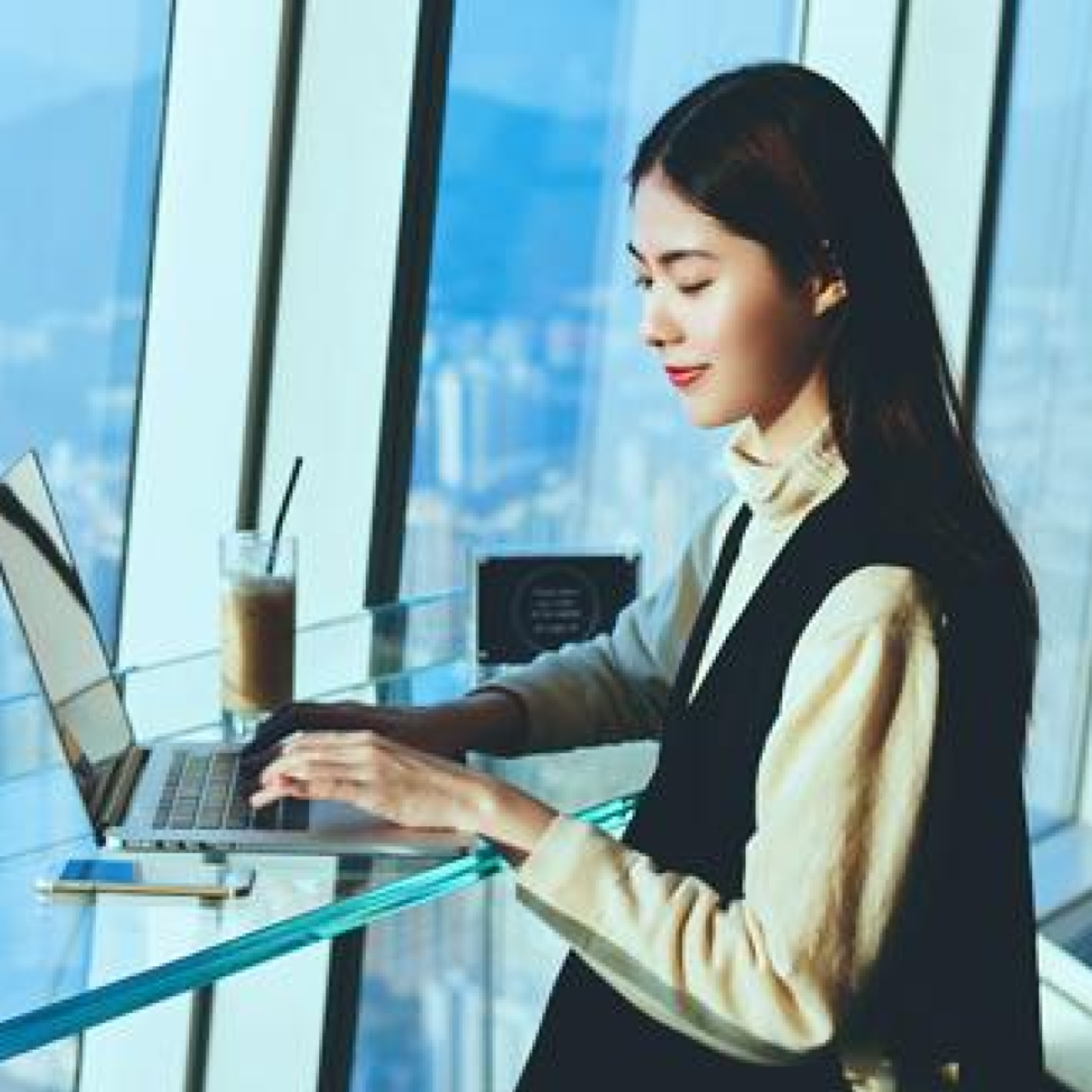 A woman typing on her laptop on a glass table by a glass window overlooking a cityscape.
